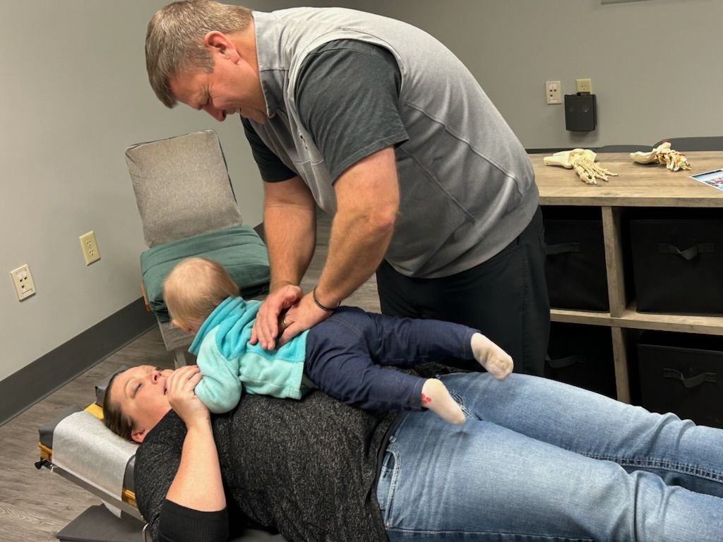 Man adjusting baby on mother's chest at chiropractic office. They are indoors.