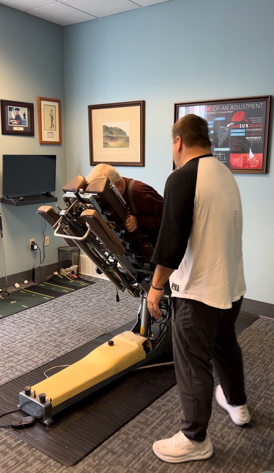 Man adjusting chiropractic table as another person leans on it in a doctor's office.