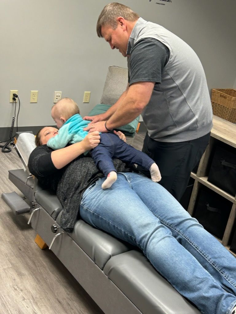 Woman lying on table holding baby as chiropractor examines. Gray table, neutral wall.