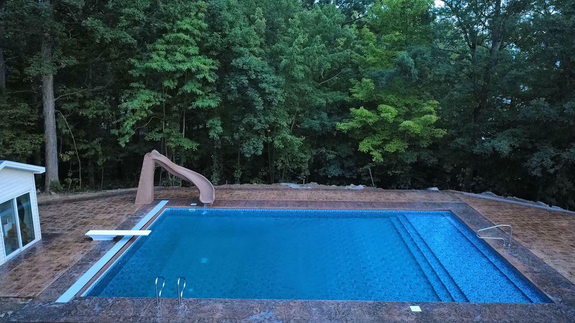 Pool with slide, diving board, and steps, surrounded by trees and a stone border. Blue water.
