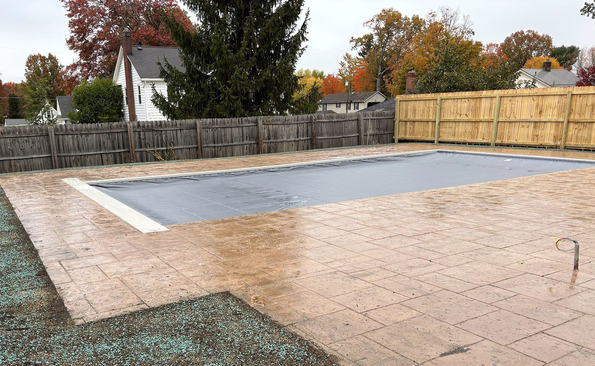 Pool covered with a tarp on a brick patio, surrounded by a fence and fall foliage.