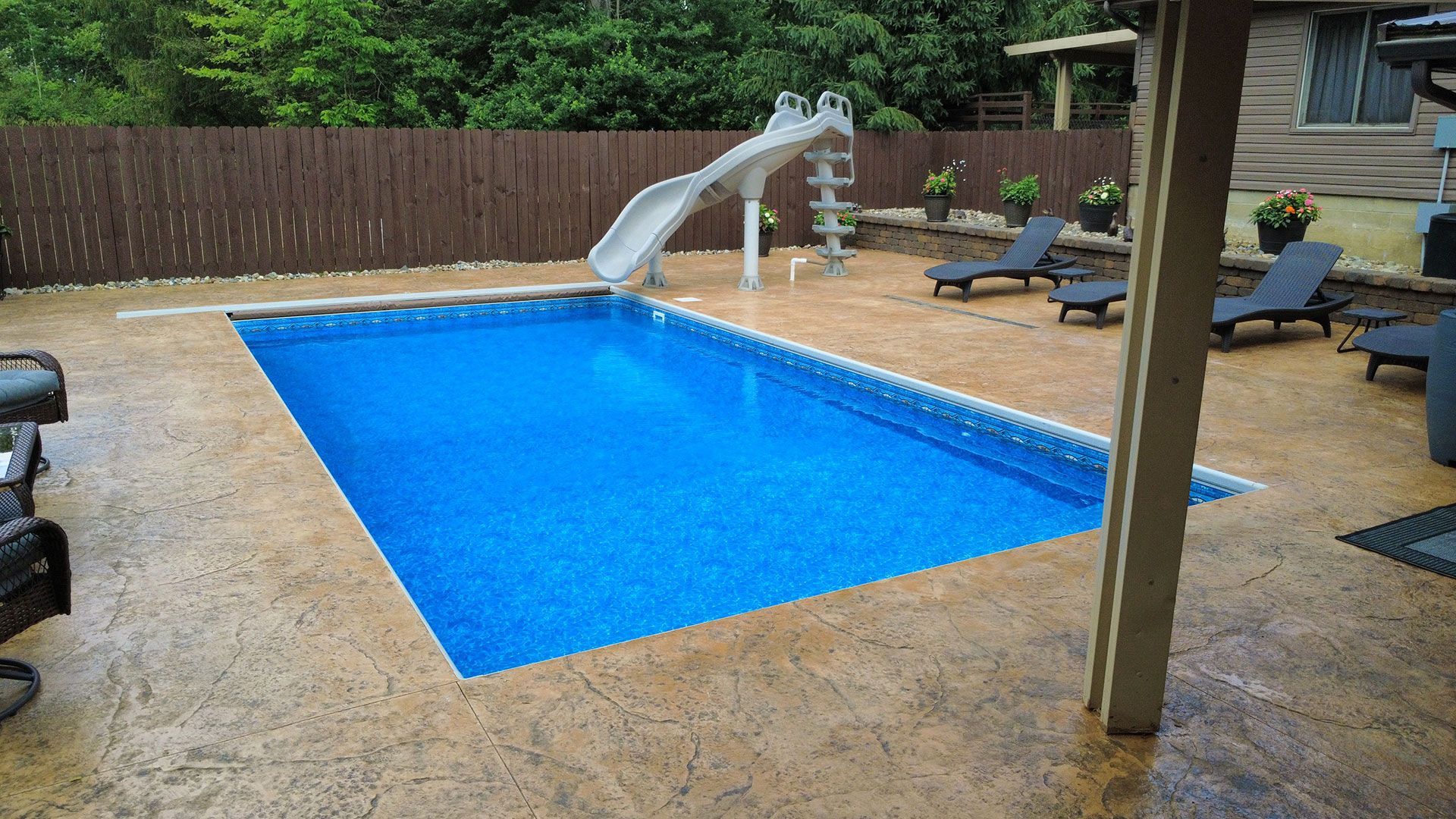 Rectangular in-ground pool with slide, surrounded by a concrete patio and a wooden fence.