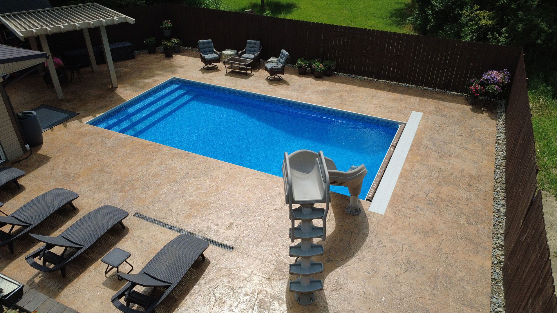Aerial view of a rectangular blue swimming pool with a slide, surrounded by a tan patio, lounge chairs, and a wooden fence.