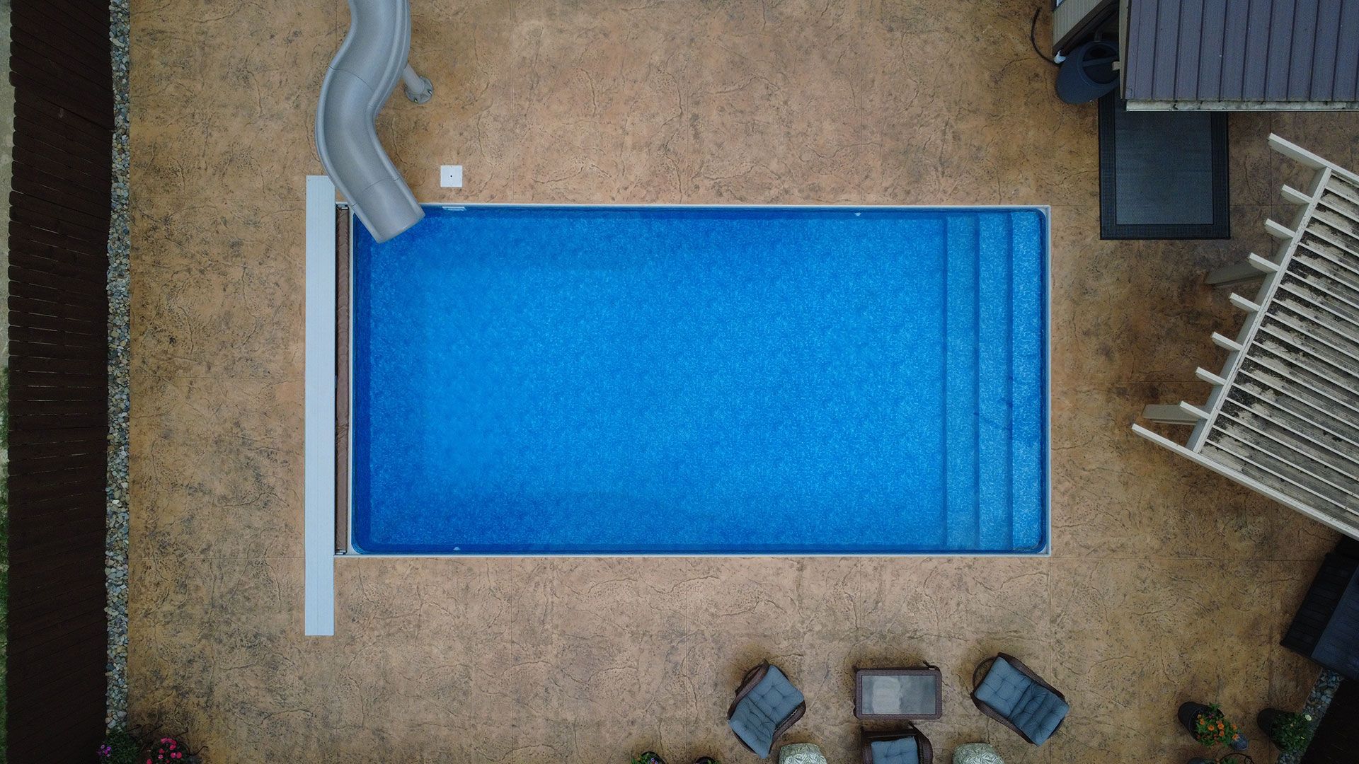 Aerial view of a rectangular blue swimming pool with a slide, steps, and surrounding patio furniture.