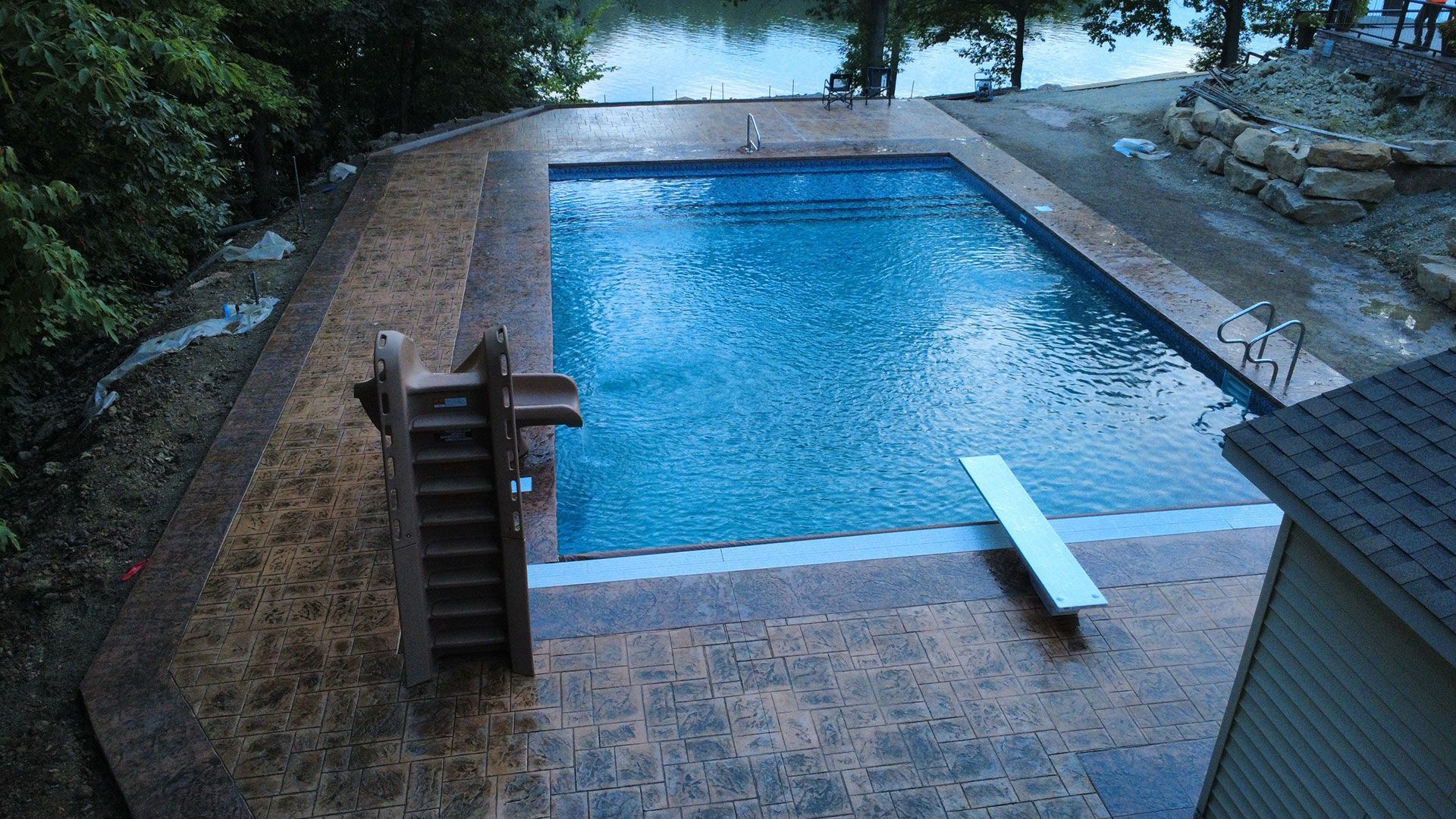 Pool with diving board and wooden steps next to body of water. Concrete surrounding the pool.