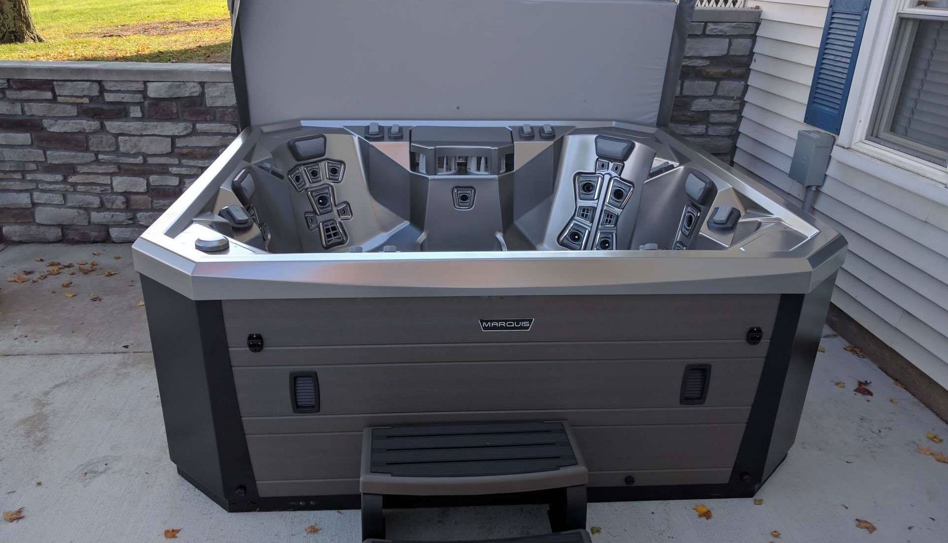 A gray and black hot tub with an open cover, steps in front, outside a house.