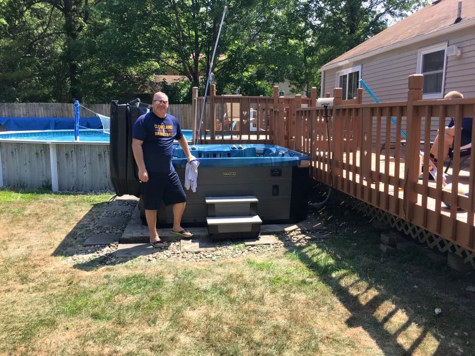 Man stands next to a new hot tub in a backyard, near a pool and a deck, sunny day.