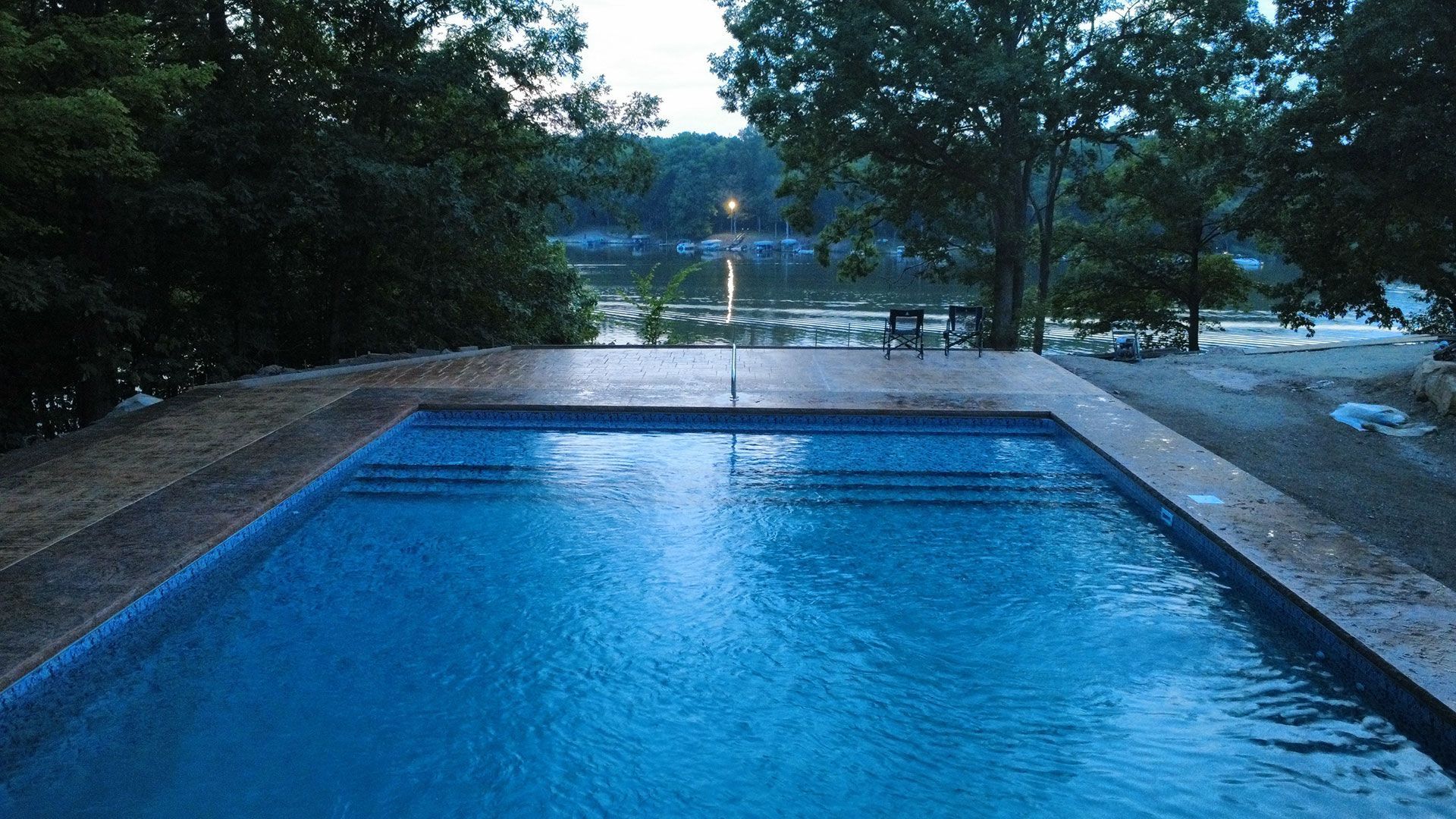 A blue swimming pool on a patio overlooking a lake surrounded by trees at twilight.