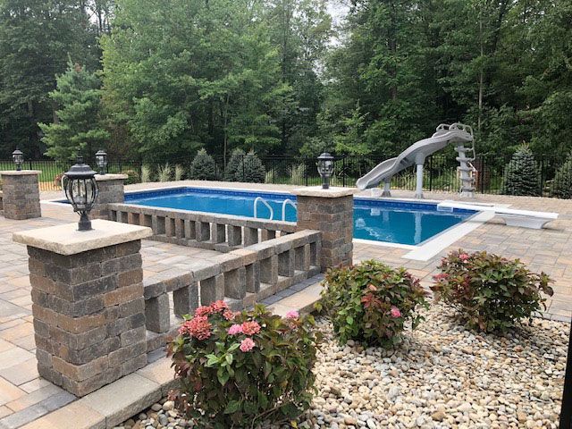 Swimming pool with slide, surrounded by a paved patio and landscaping. Dark brown brick pillars with lights.