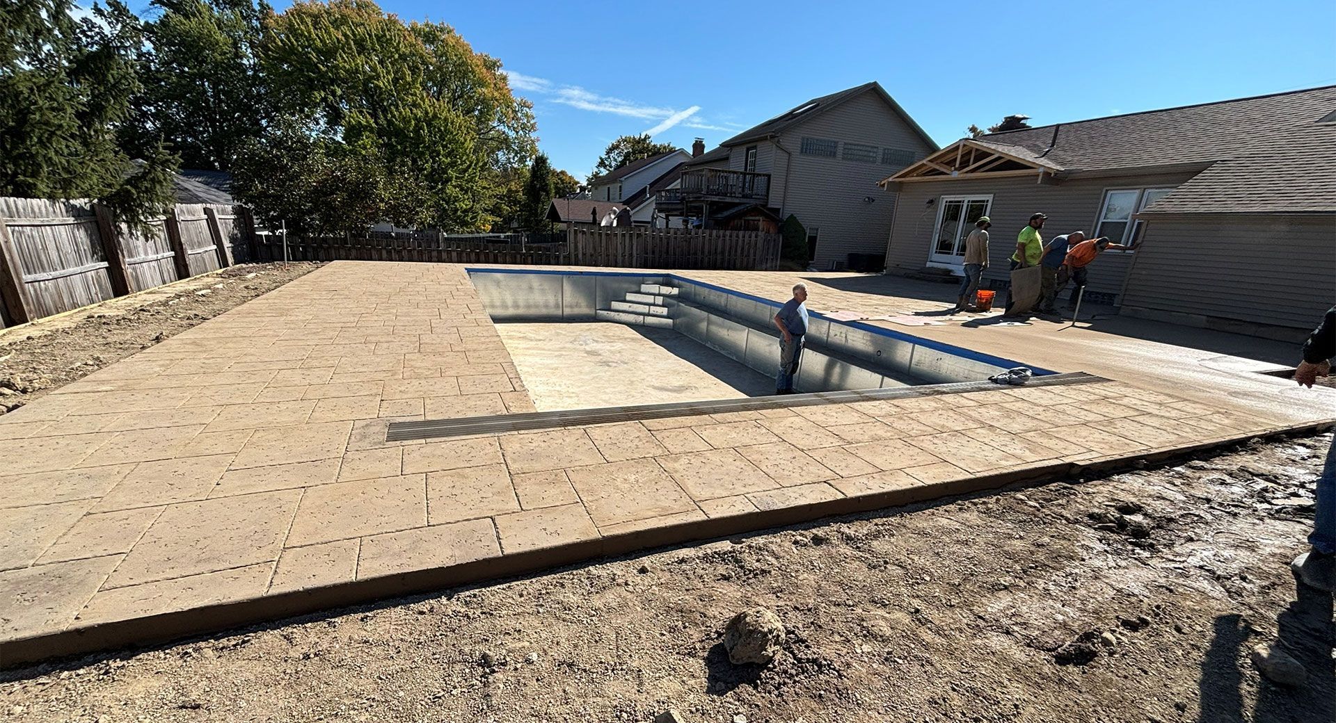 Pool under construction in a yard. Workers are present. Soil and paving stones surround the pool frame. Sunny day.