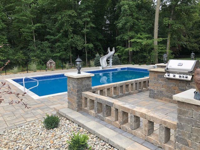 A person relaxes in a square hot tub located on a covered patio with a stone fireplace and view of a garden.