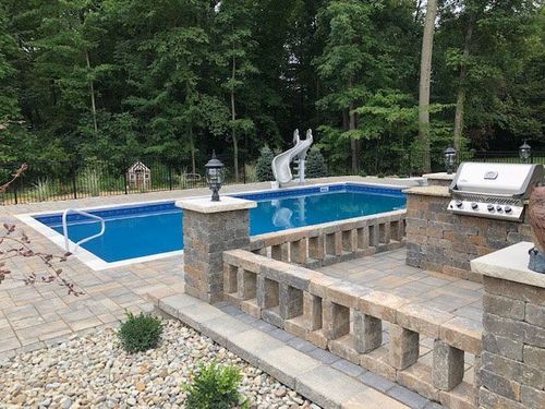 A person relaxes in a square hot tub located on a covered patio with a stone fireplace and view of a garden.