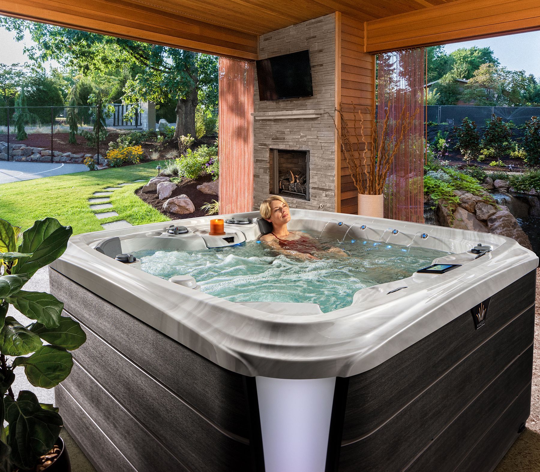 A person relaxes in a square hot tub located on a covered patio with a stone fireplace and view of a garden.