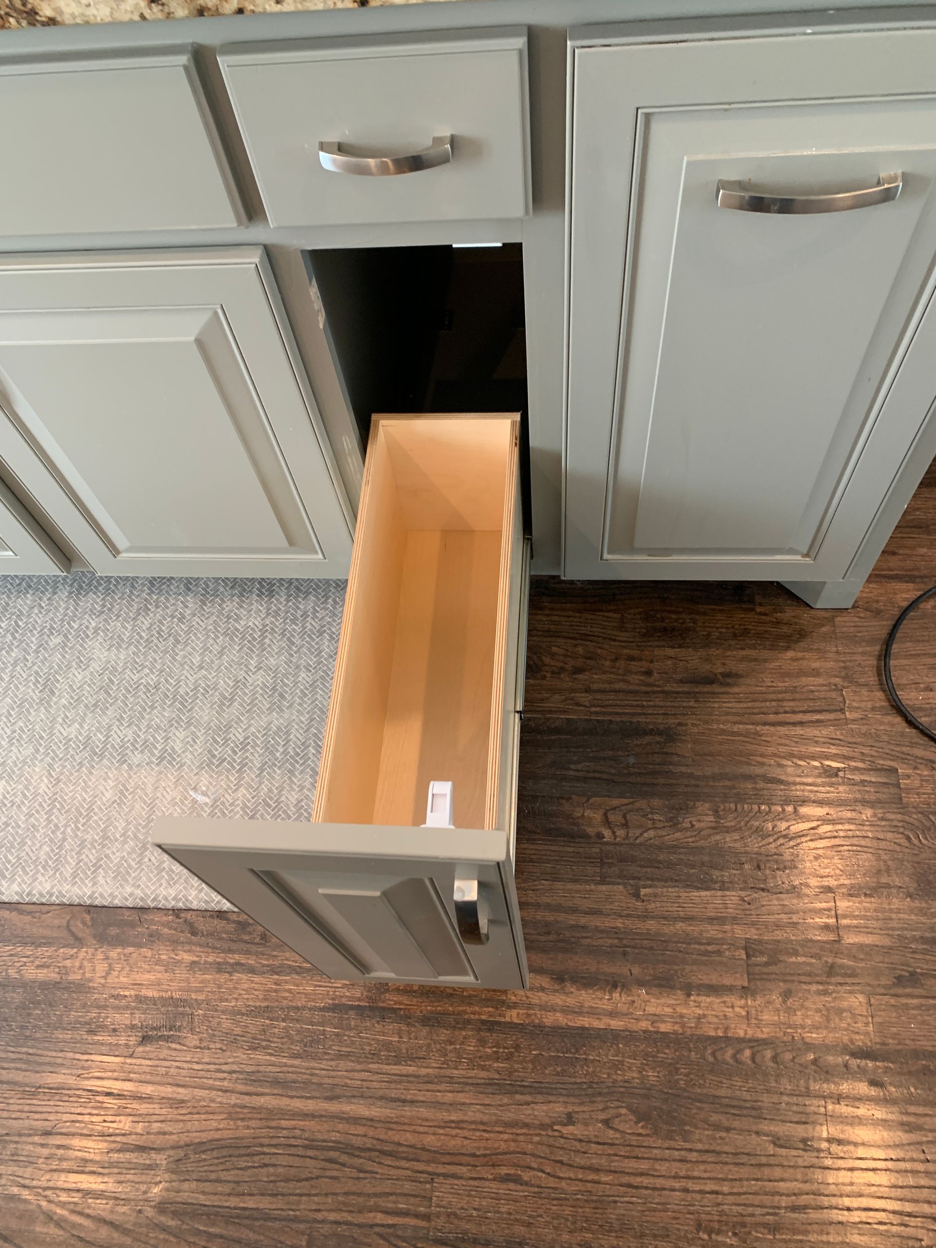 A gray kitchen cabinet with a partially open pull-out drawer. Wooden floors and a light gray rug are visible.