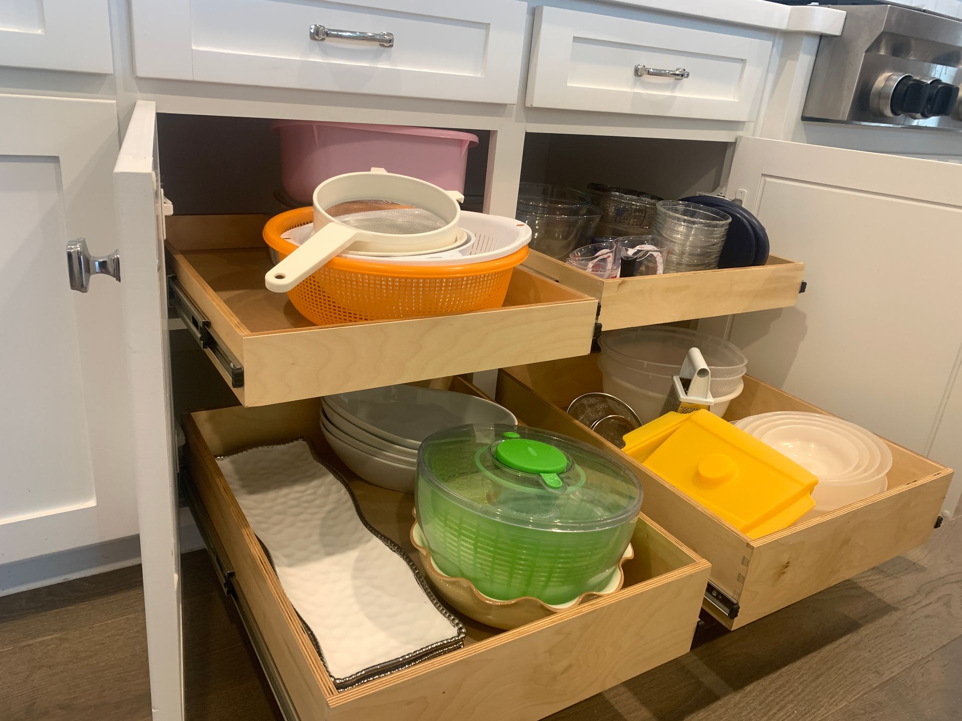 Pull-out kitchen cabinet drawers with dishware and utensils, including colanders, plates, and a salad spinner.