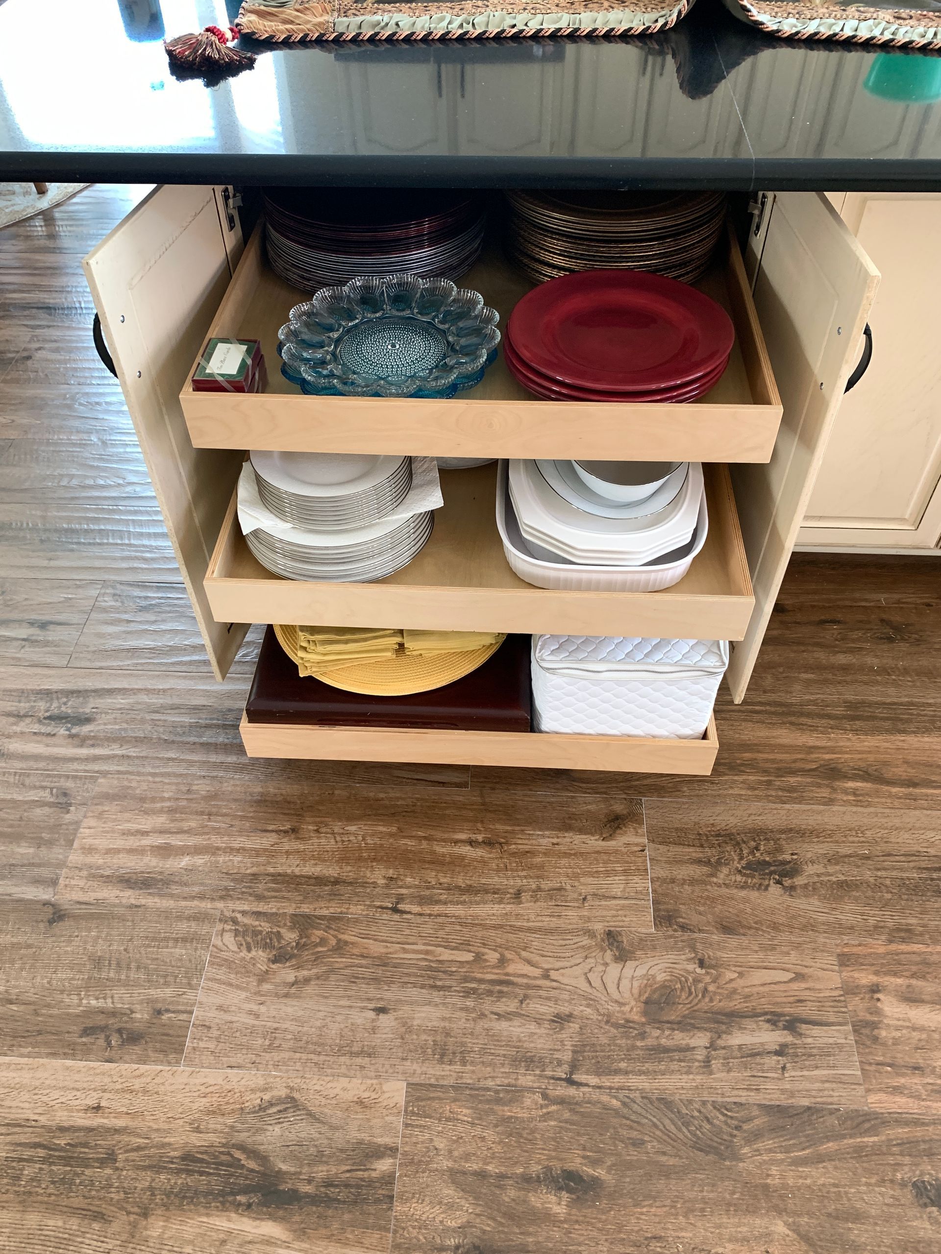 Pull-out shelves in a kitchen island hold stacks of plates and bowls of various colors, on a wood-look floor.