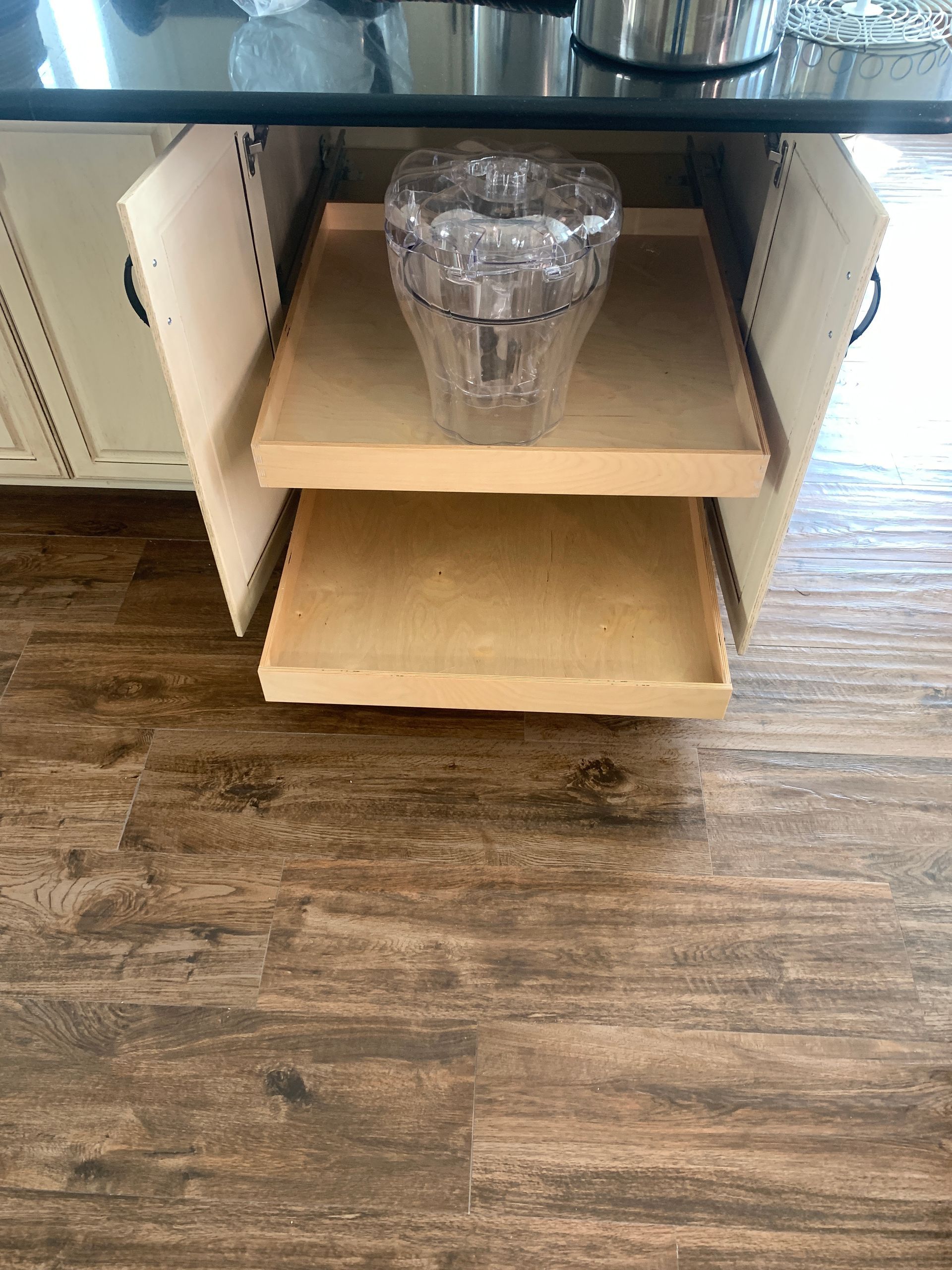 Two pull-out shelves in a kitchen cabinet, holding a clear glass vase. Wooden floor.