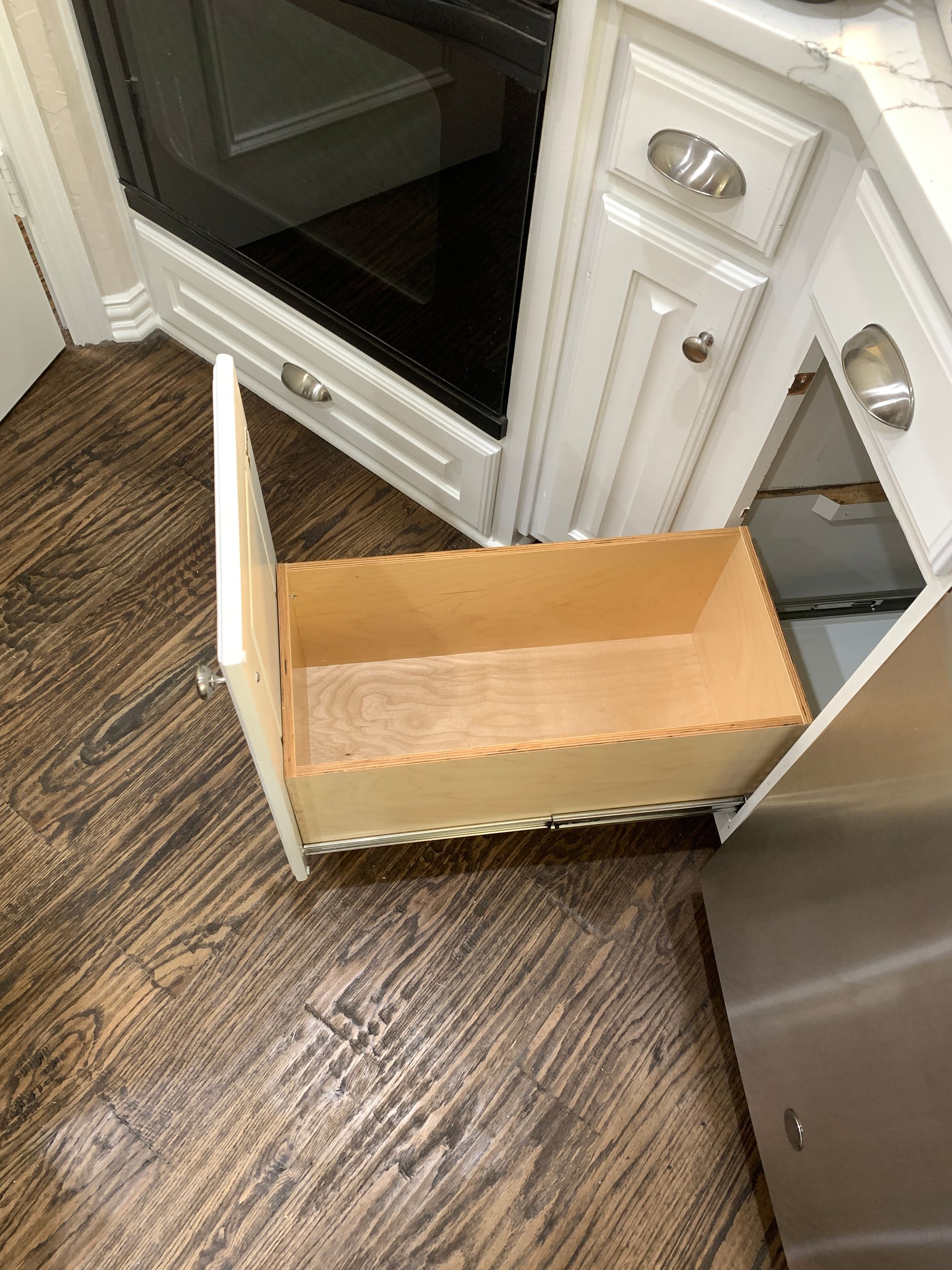 Open kitchen drawer in a white cabinet; light wood drawer box, wood floor, stainless steel oven.