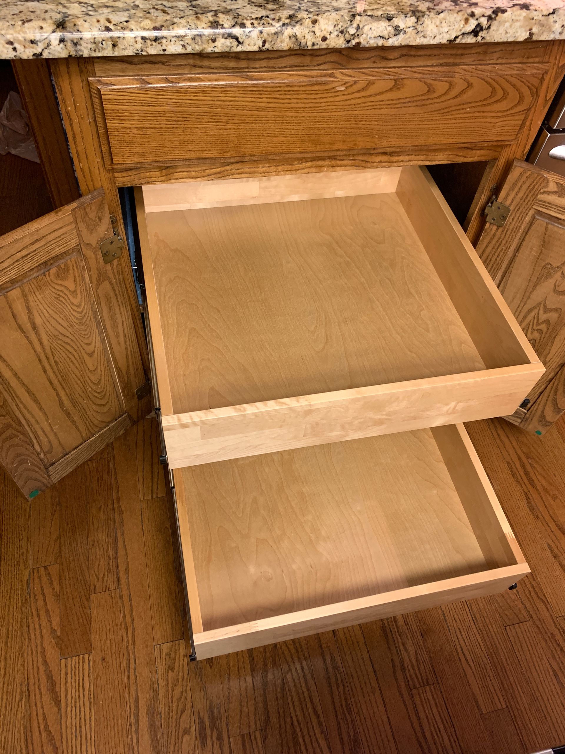Two wooden kitchen drawers pulled out from a light brown cabinet, with cabinet doors on the sides, on a hardwood floor.