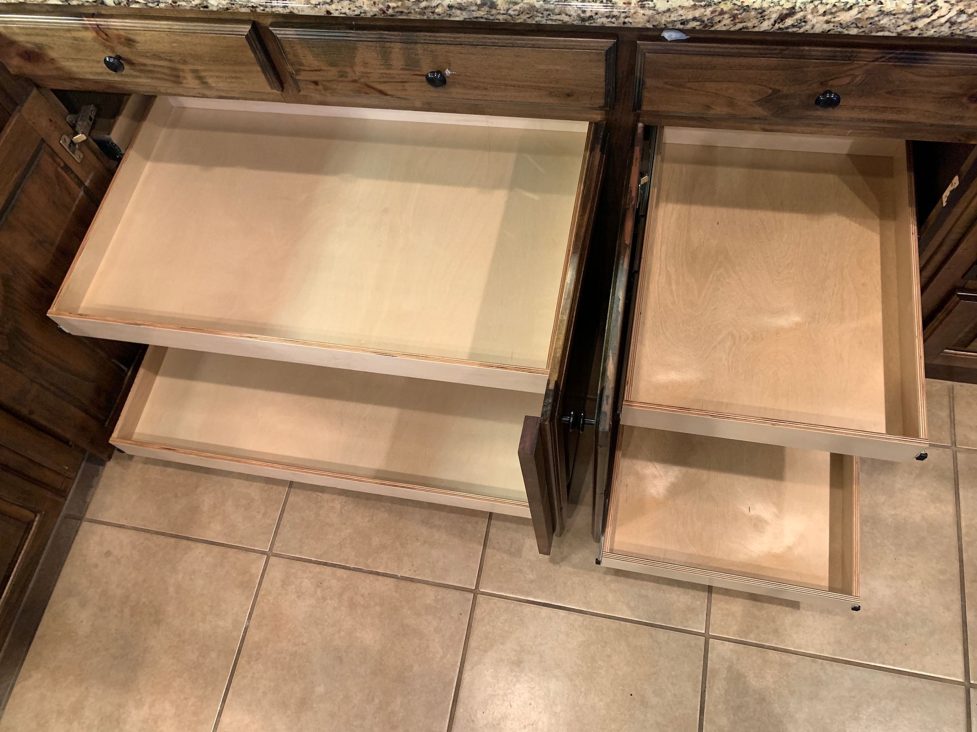 Open kitchen drawers; wooden cabinets with beige tile floor.