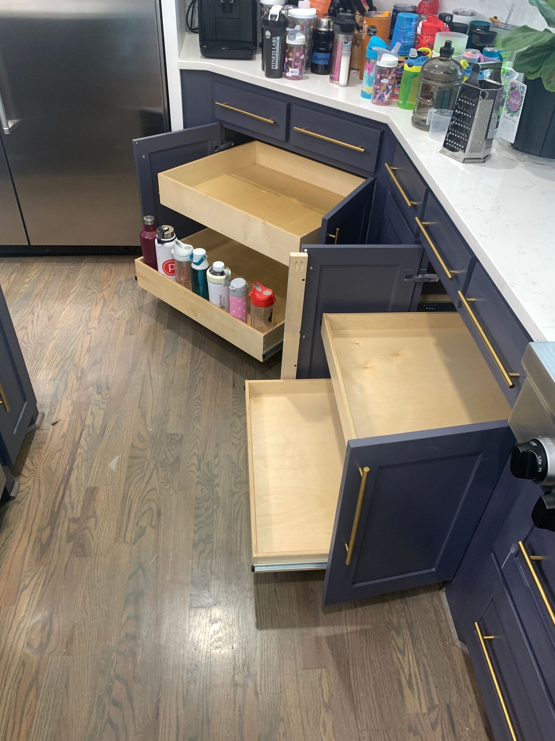 Dark blue kitchen cabinets with pull-out shelves, some open, holding spices and bottles. Wooden floor.