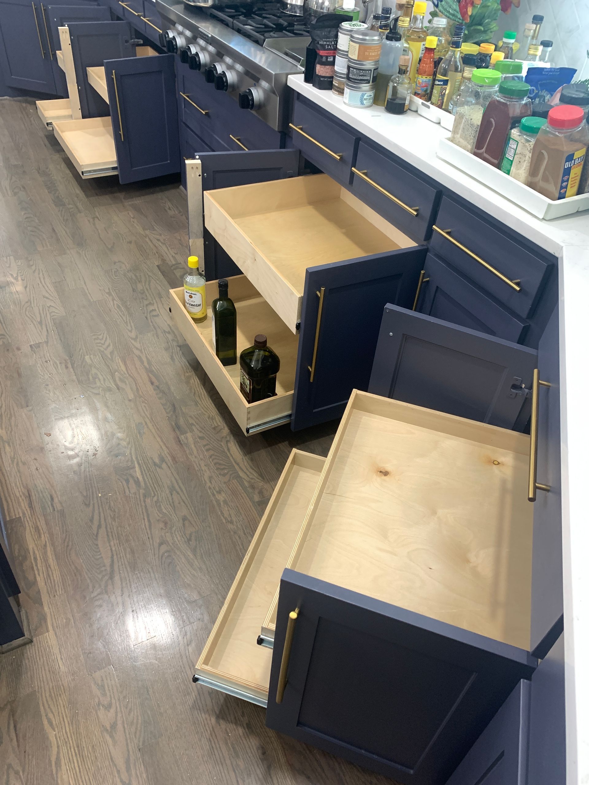 Dark blue kitchen cabinets with gold hardware, open to reveal pull-out drawers on a wood floor.