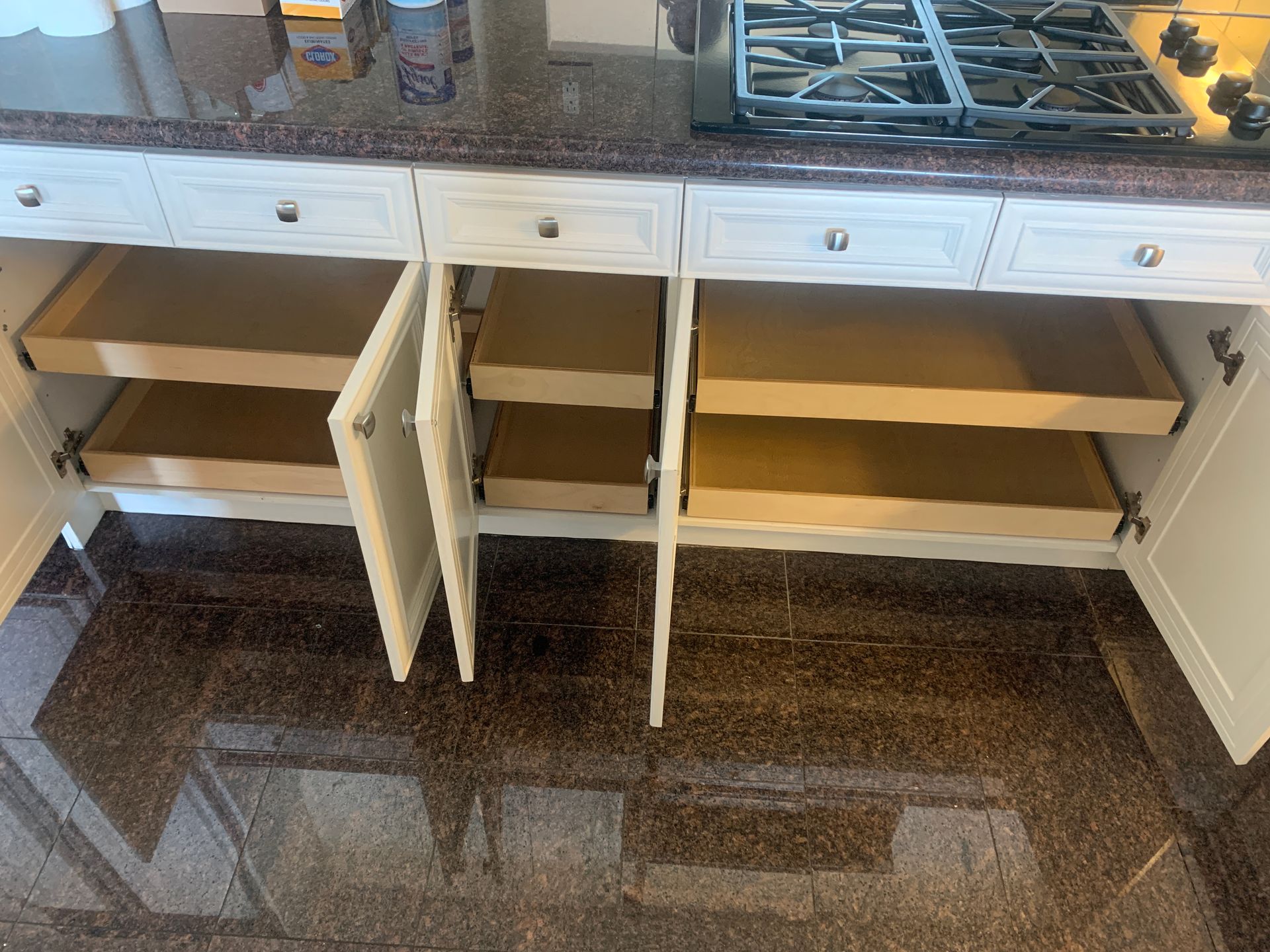 Kitchen cabinets open, revealing pull-out drawers. Beige cabinets and drawers. Brown granite countertop.