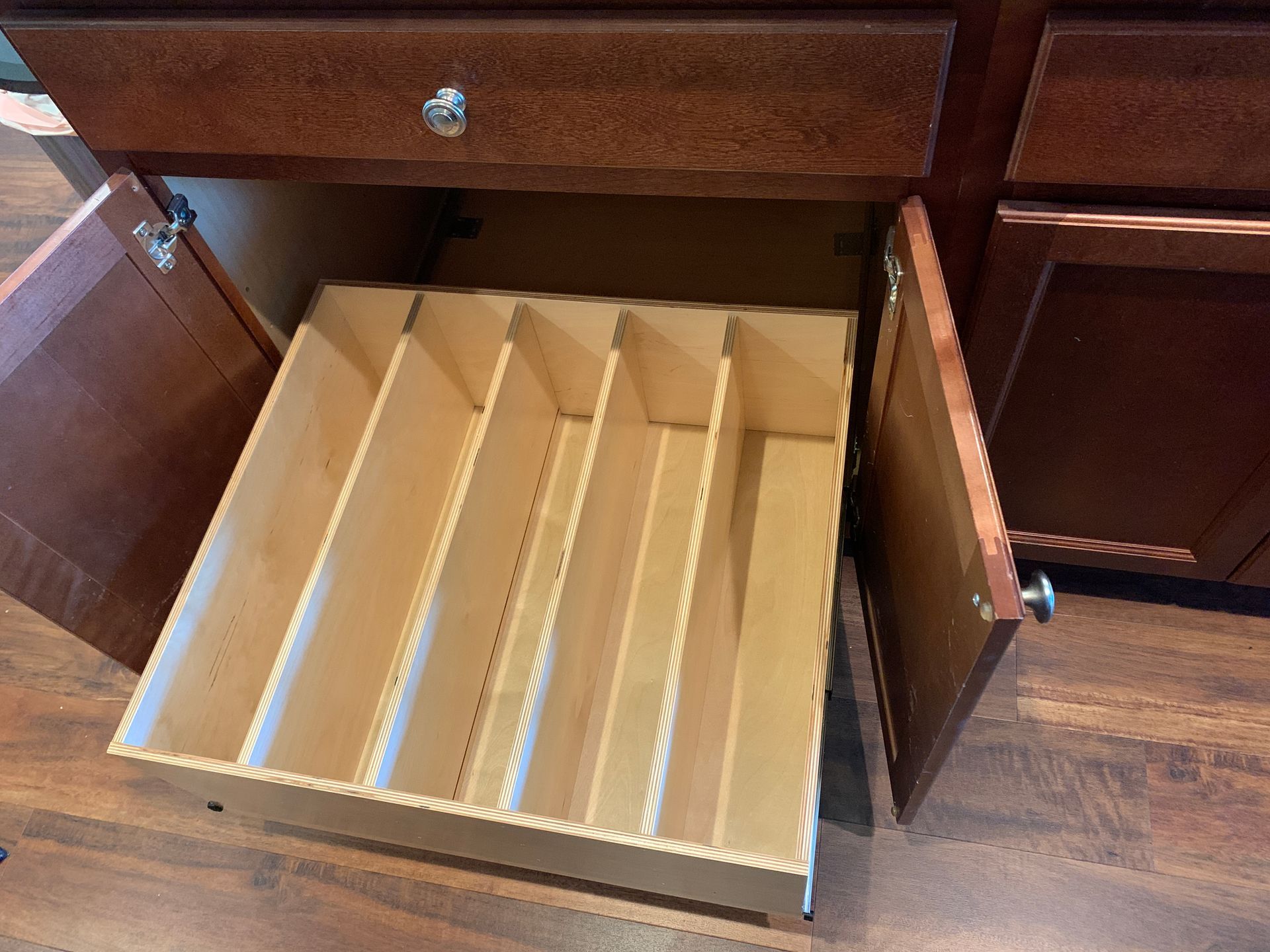 A kitchen cabinet with a pull-out wooden drawer containing several dividers, open on a wooden floor.