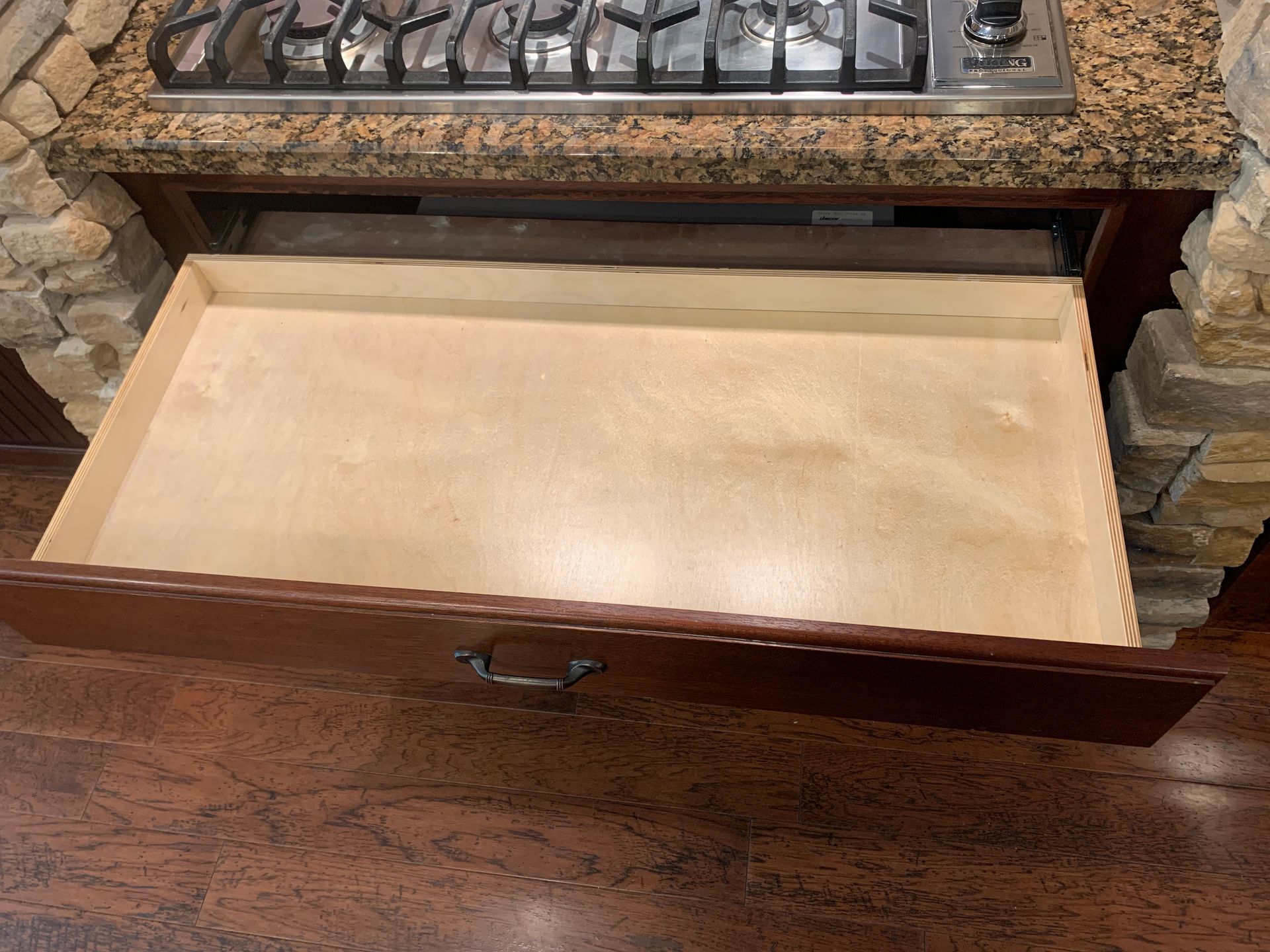 Open drawer under a stove, beige interior, brown wood exterior and counter, stone wall.