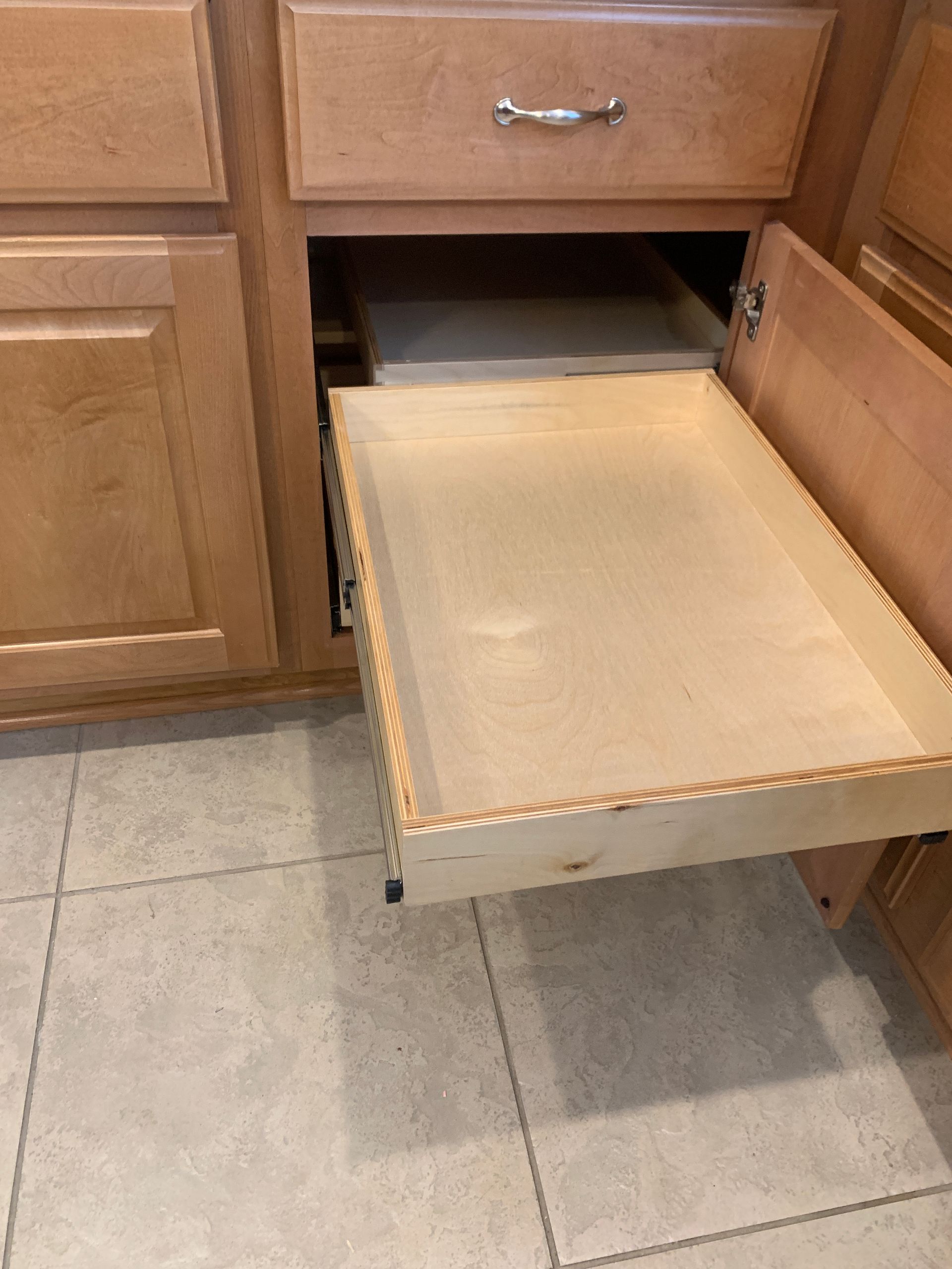 Open wooden kitchen cabinet with a pull-out drawer extended. Light wood cabinets and beige tile floor.