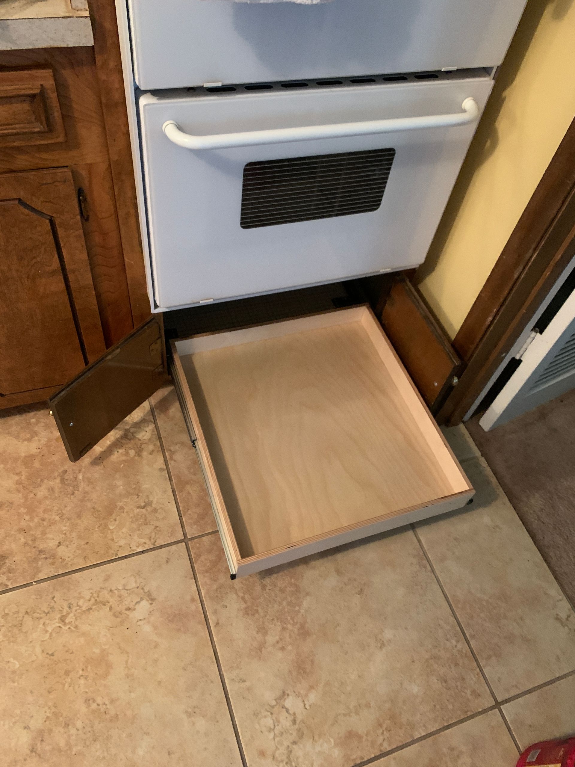 A pull-out drawer beneath a white oven, on a tile floor in a kitchen.