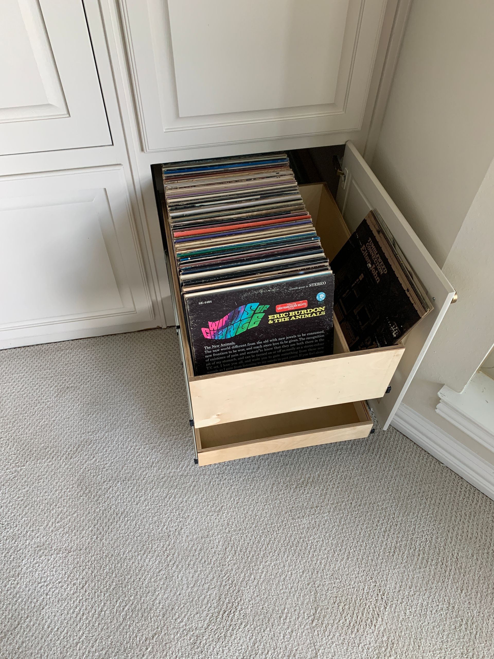 Wooden drawer holding vinyl records, inside a white cabinet, on a gray carpet.
