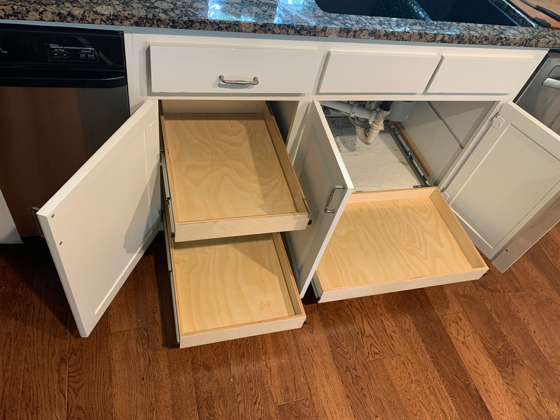 Kitchen cabinet with open doors and pull-out drawers; dishwasher on the left, wood floor.