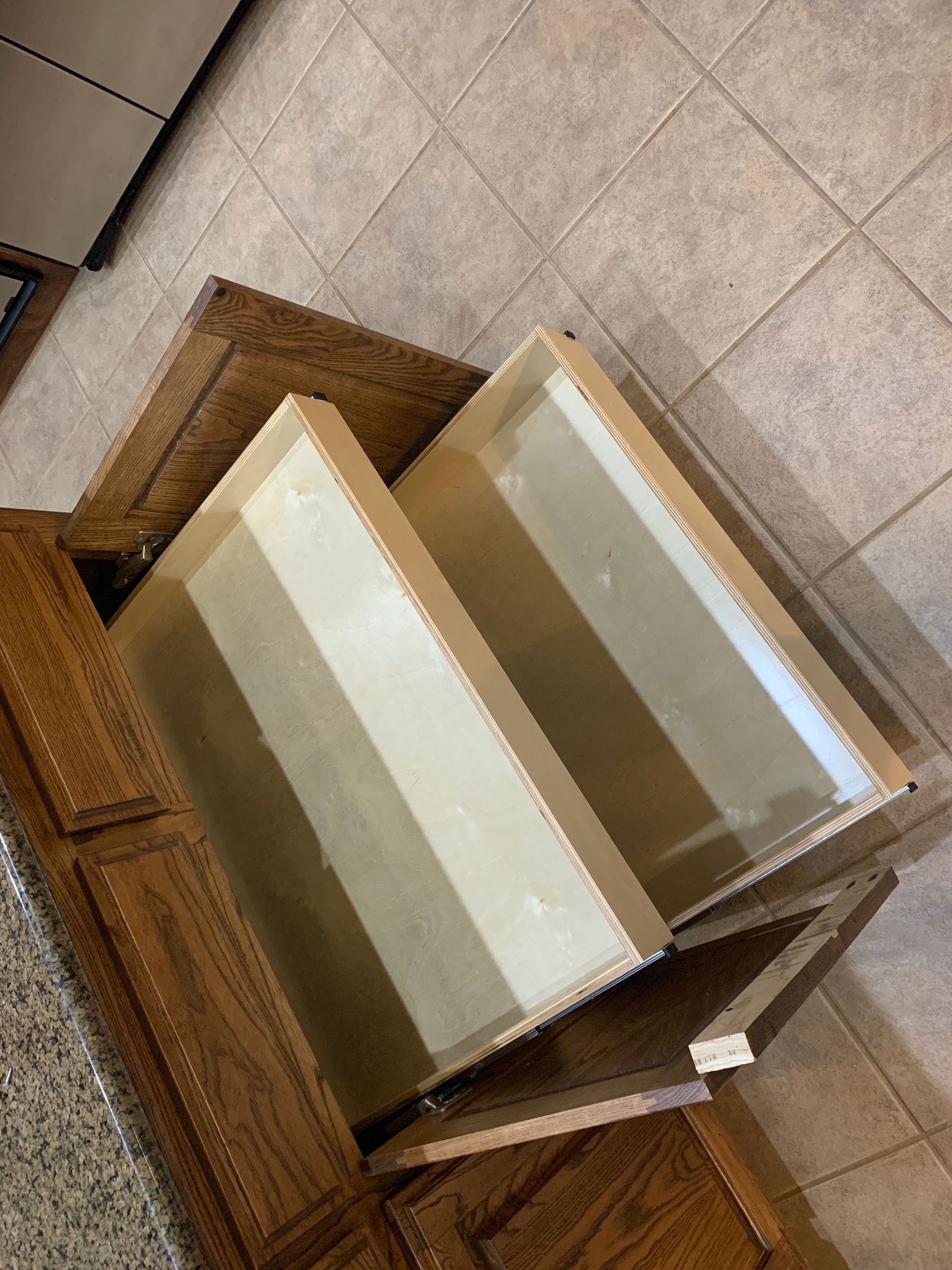 Wooden kitchen cabinet with two drawers partially pulled out. Light brown wood and beige tile floor.