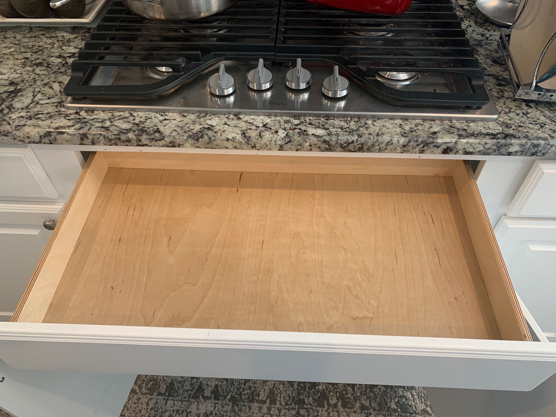 Open, empty kitchen drawer beneath a stovetop. The drawer is wood-toned and white.