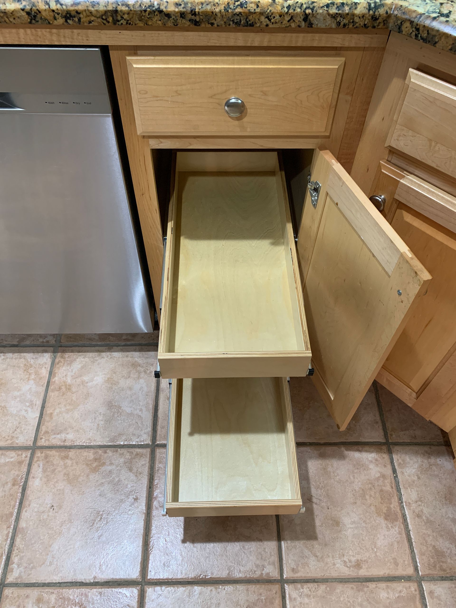 A kitchen cabinet with a pull-out spice rack and partially open door; light wood and tile floor.