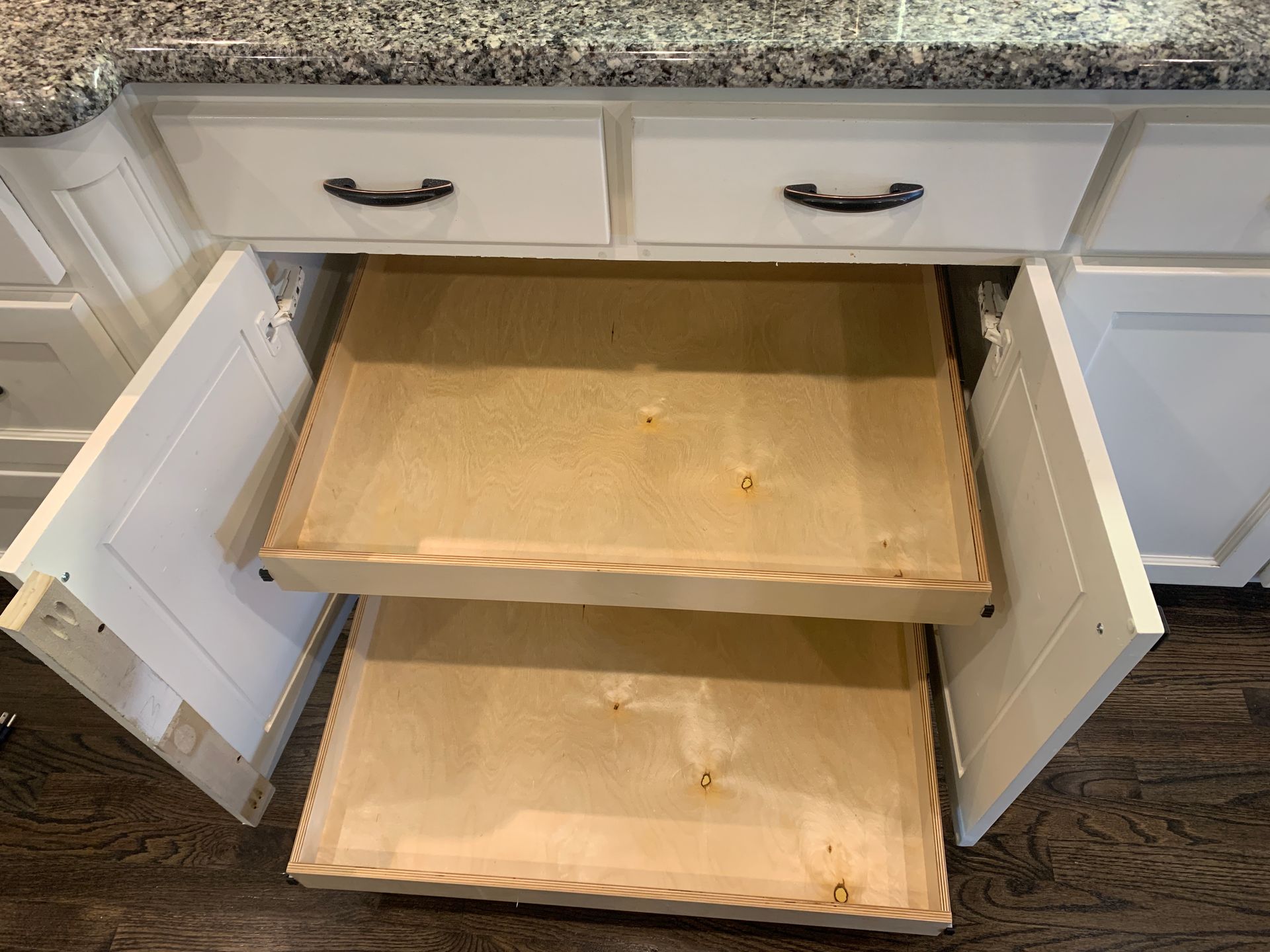 Open white kitchen cabinet revealing two wooden pull-out shelves under a granite countertop.