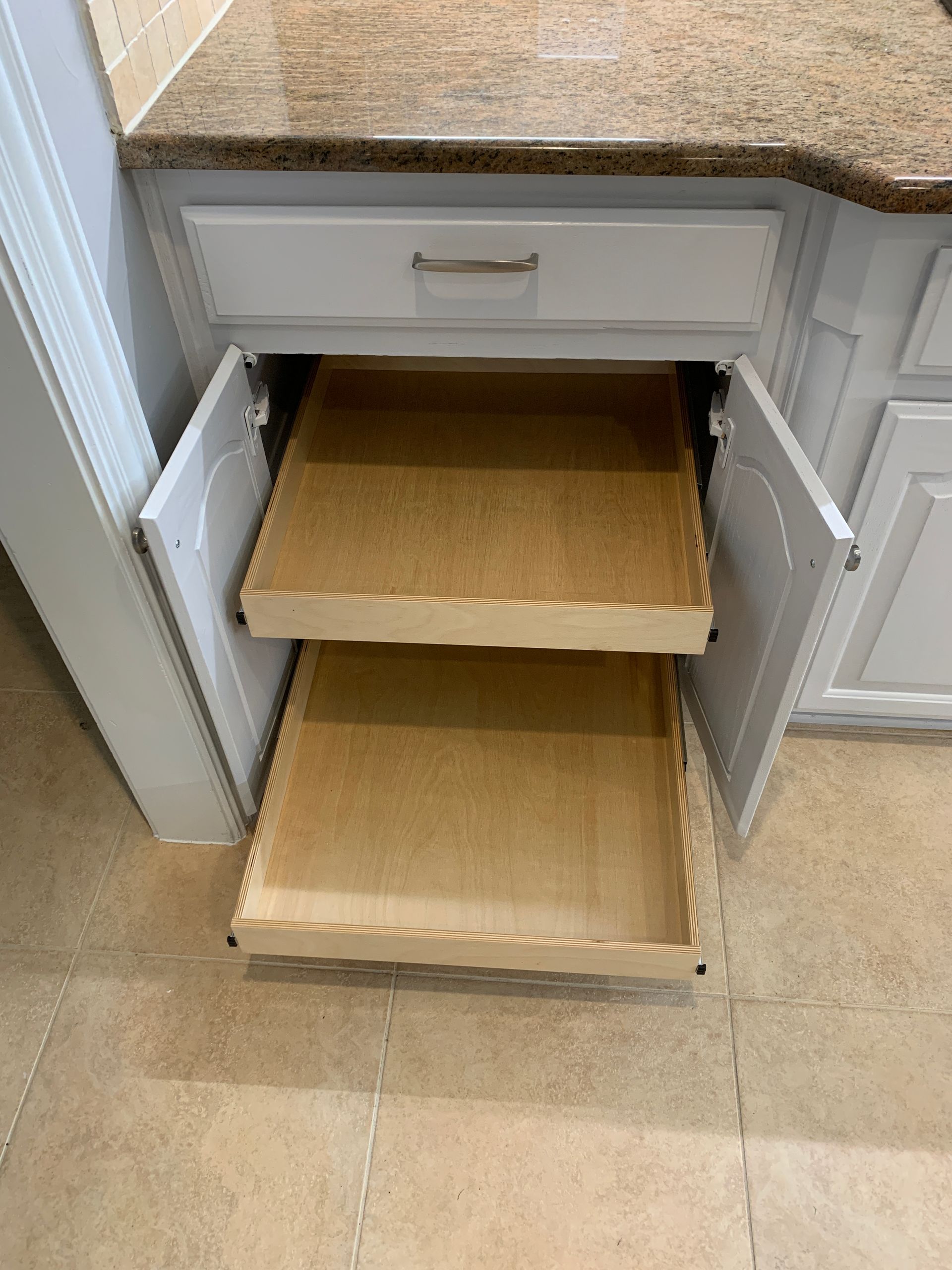 White kitchen cabinet with pull-out shelves. Granite countertop. Open doors revealing two wooden shelves.