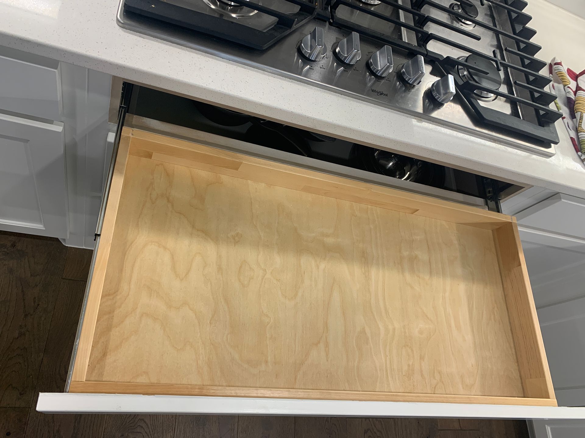 Open, empty drawer beneath a stovetop in a kitchen. Light wood interior, white trim.