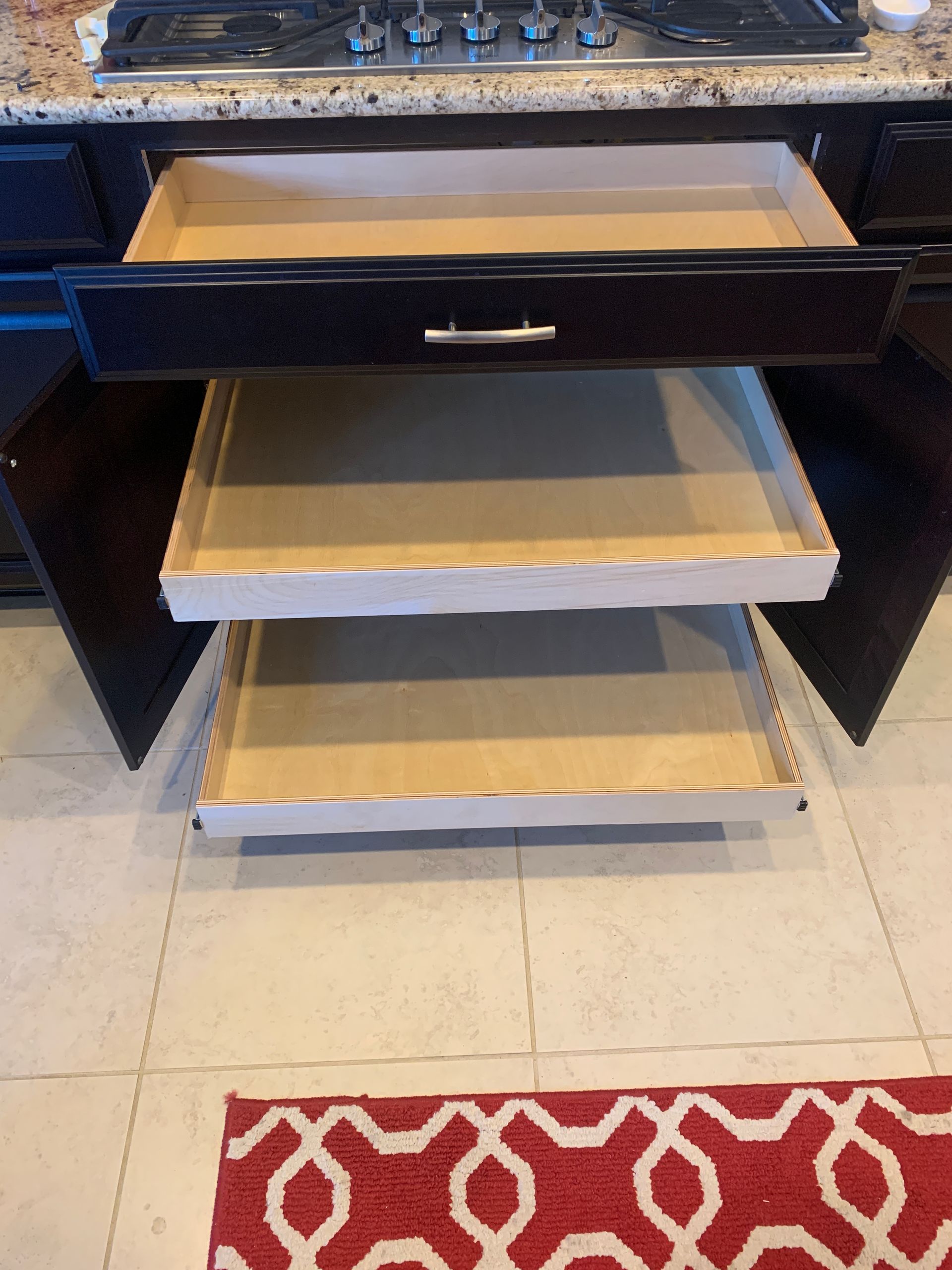 Dark cabinet with three pull-out drawers, two open, set in a kitchen. Beige tiled floor with a red rug.