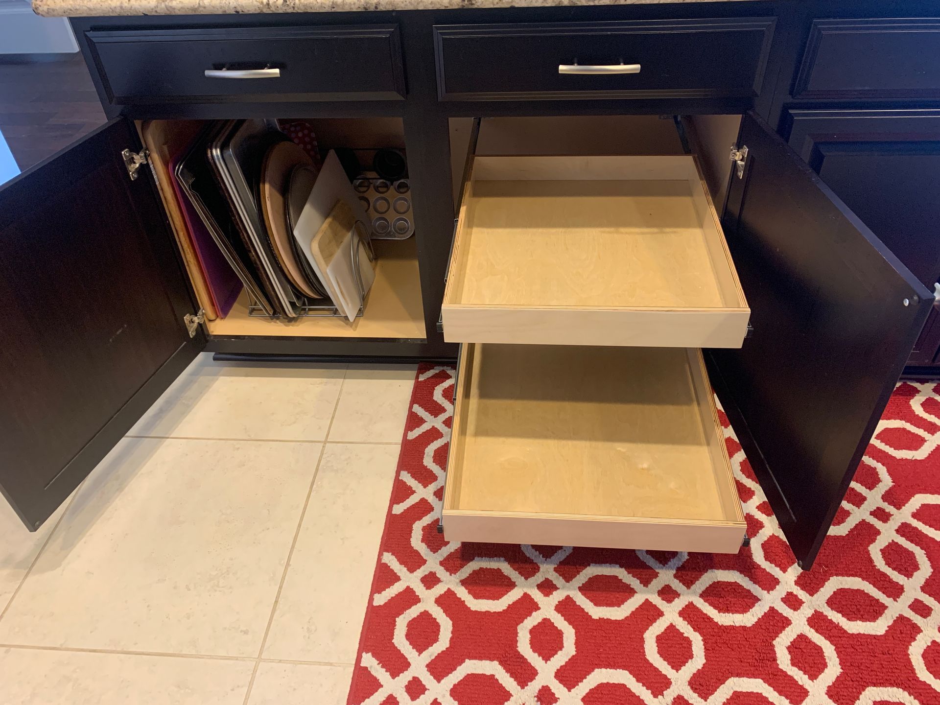 Kitchen cabinet open, with pull-out drawers, containing baking pans. Red patterned rug visible.