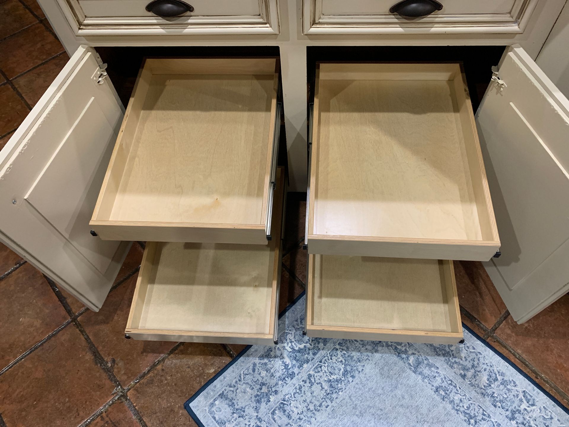Open kitchen cabinet with four wooden pull-out drawers.  Beige cabinet and dark hardware,  brown floor.
