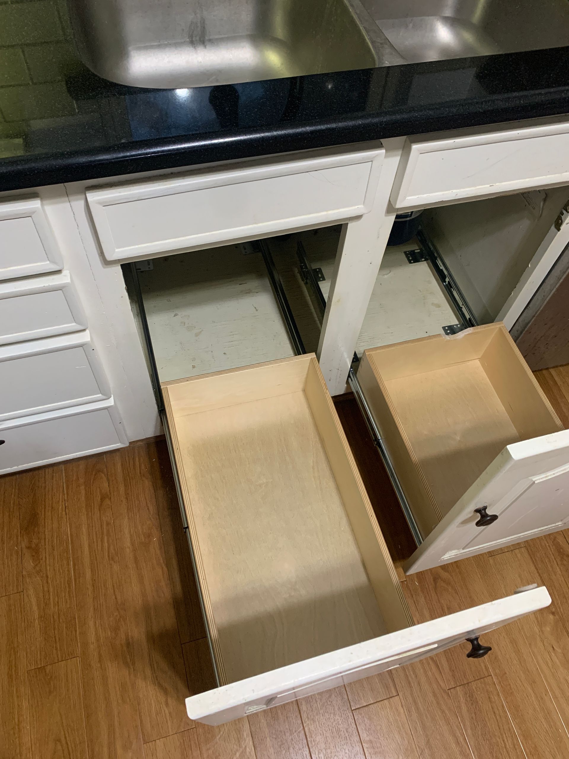 Open kitchen drawers below a sink. Wood drawers with metal slides, white cabinets, and black countertop.