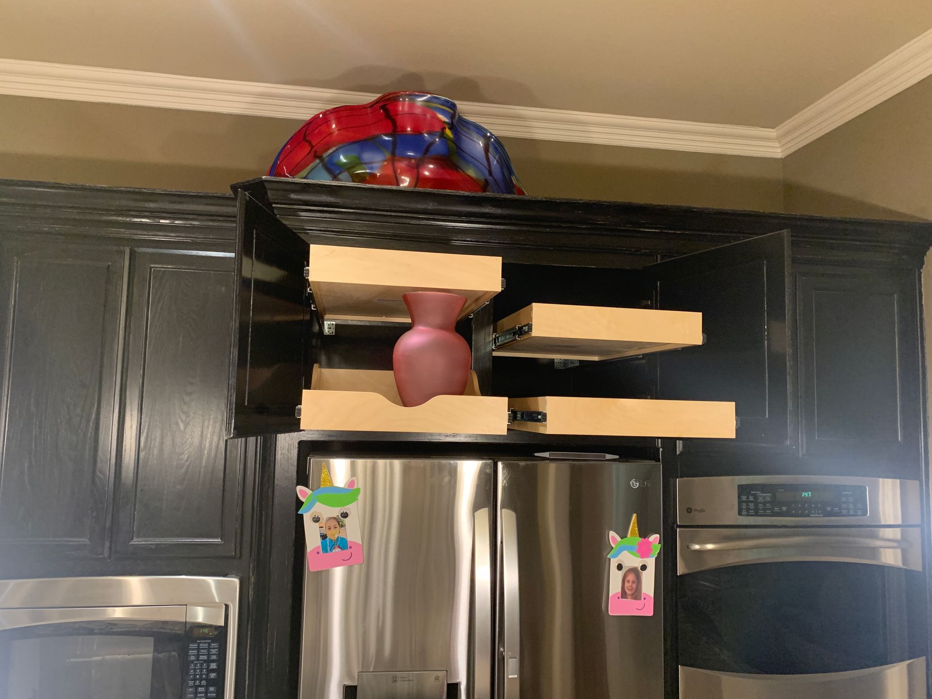 Kitchen cabinets with pull-out shelves and a vase, above a refrigerator with unicorn drawings.