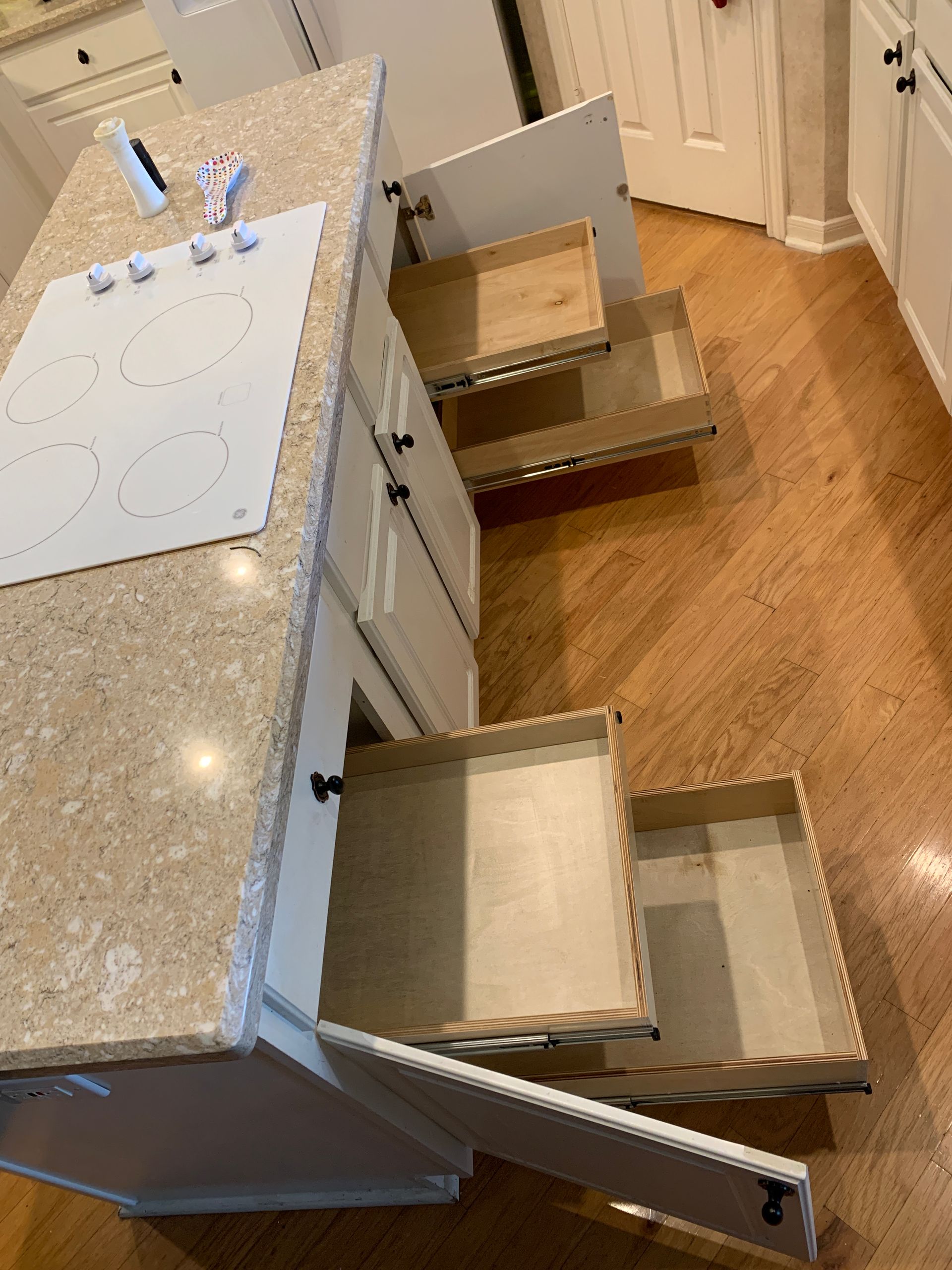 Kitchen island with open drawers and stove top. Light-colored cabinets, countertop, and hardwood floor.