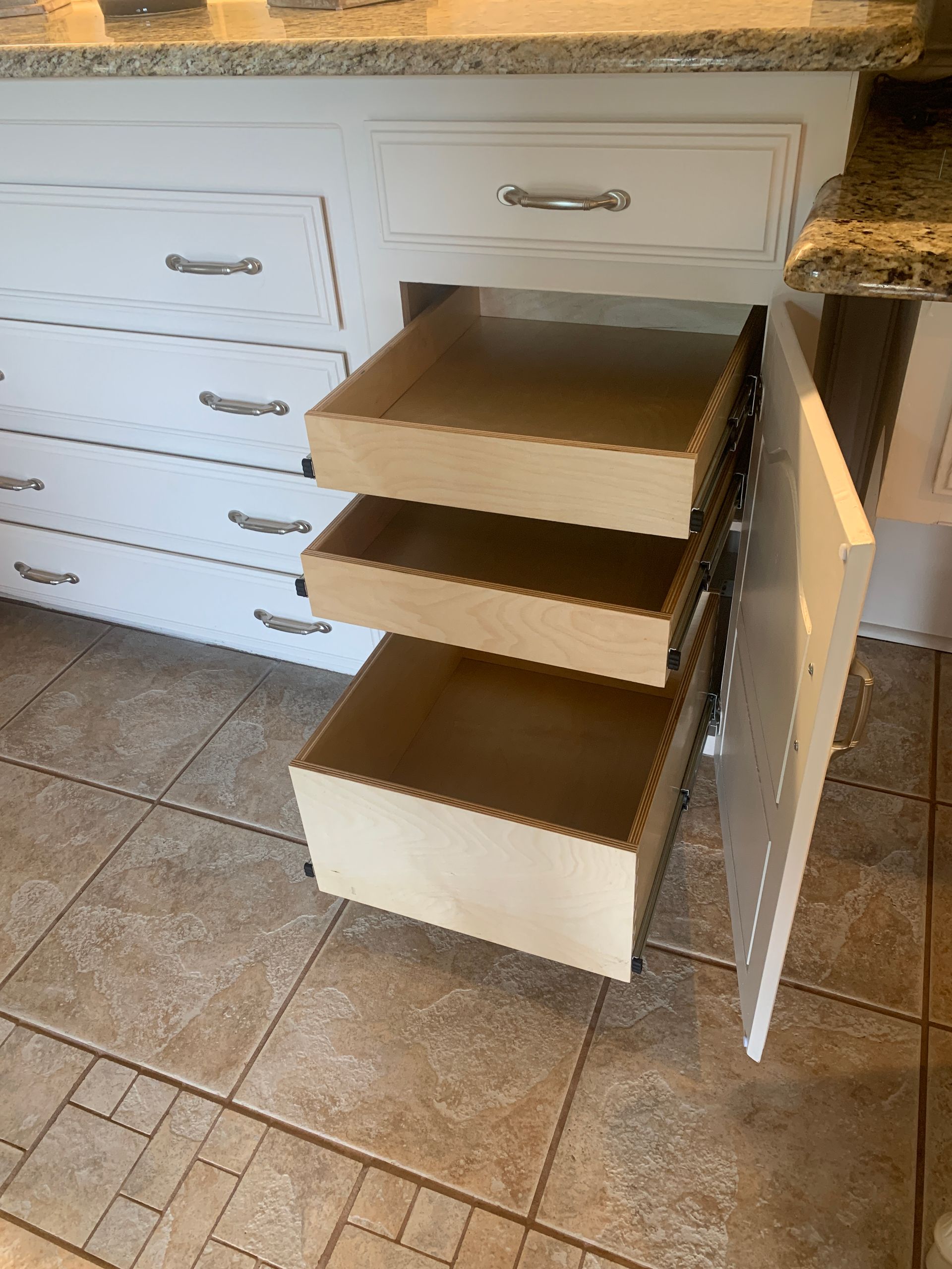White kitchen cabinet with three pull-out drawers; open. Light wood inside, light granite countertop.
