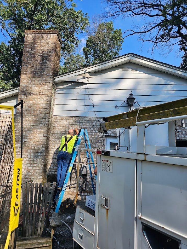 A person on a ladder repairs siding on a house next to a brick chimney and white utility truck.