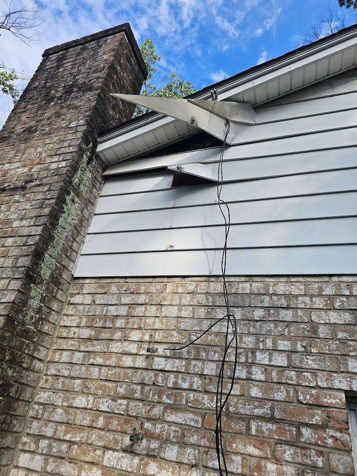 Damaged siding and roof near a brick chimney, with cracked siding and exposed structure.