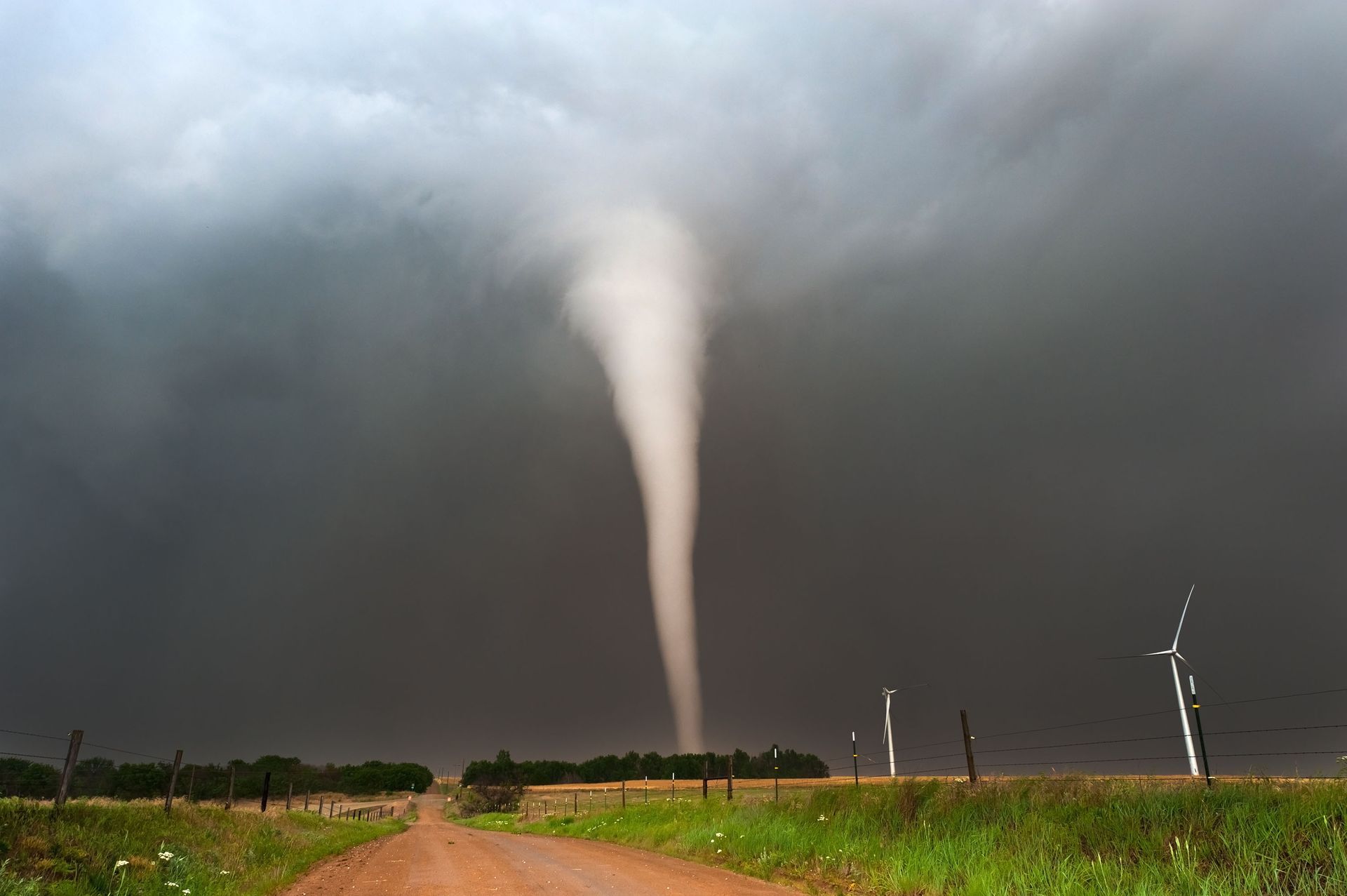 Tornado funnel descends from dark clouds over a dirt road and field with three wind turbines.