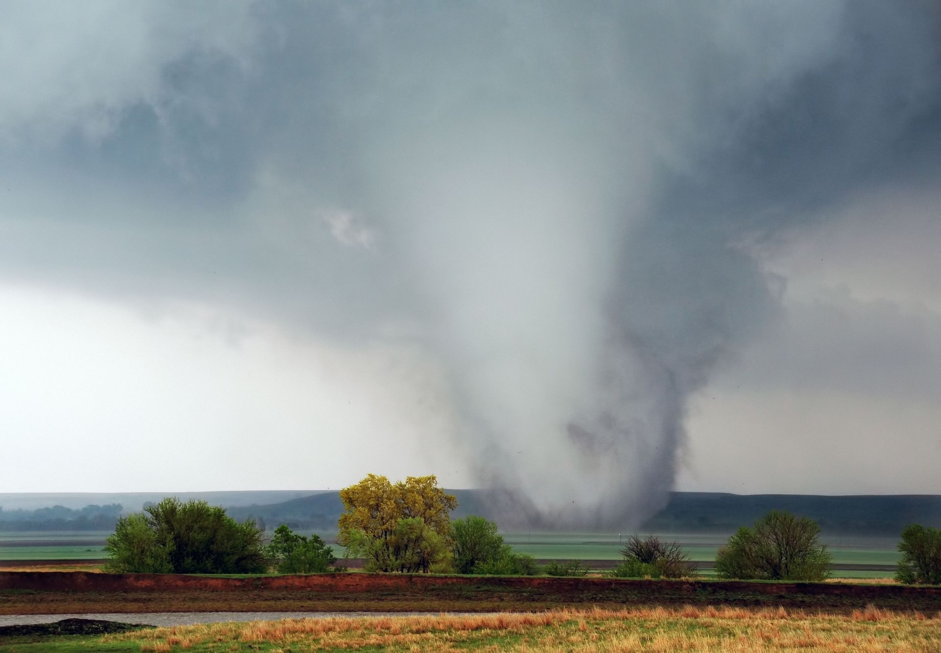 Tornado descends from dark clouds over a rural landscape, near trees and farmland.